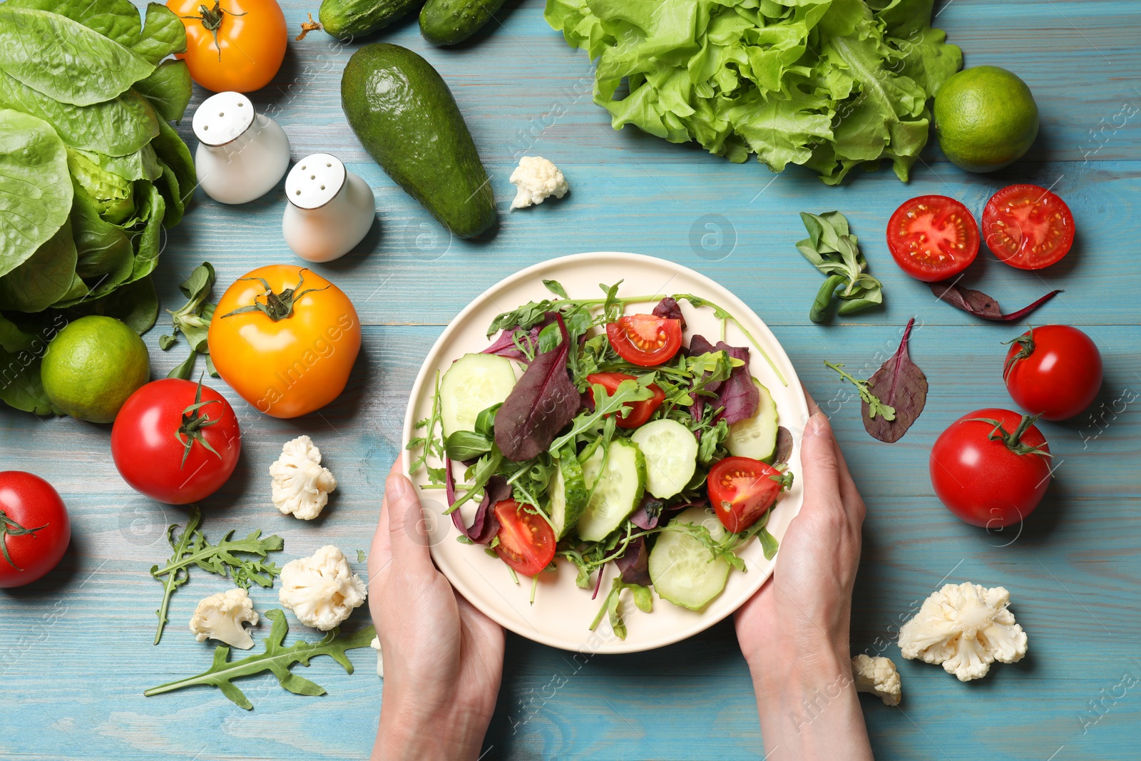 Photo of Healthy vegetarian food. Woman holding plate of salad at light blue wooden table with vegetables, top view