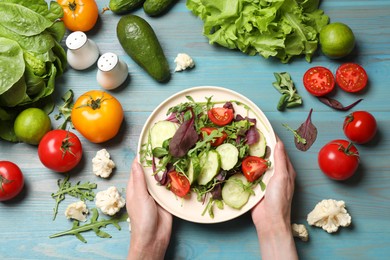 Photo of Healthy vegetarian food. Woman holding plate of salad at light blue wooden table with vegetables, top view