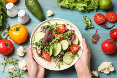 Photo of Healthy vegetarian food. Woman holding plate of salad at light blue wooden table with vegetables, top view