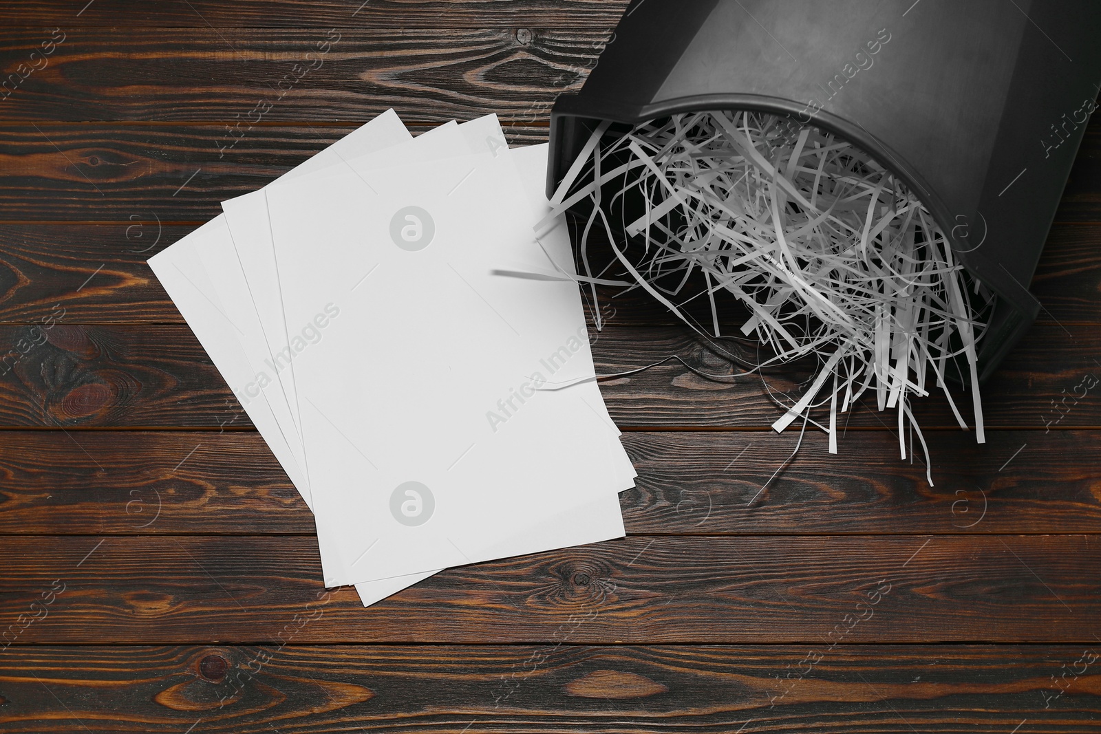 Photo of Black bin with paper strips and sheets on wooden table, flat lay