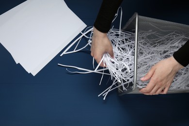 Photo of Woman putting shredded paper strips into trash bin at blue wooden table, top view