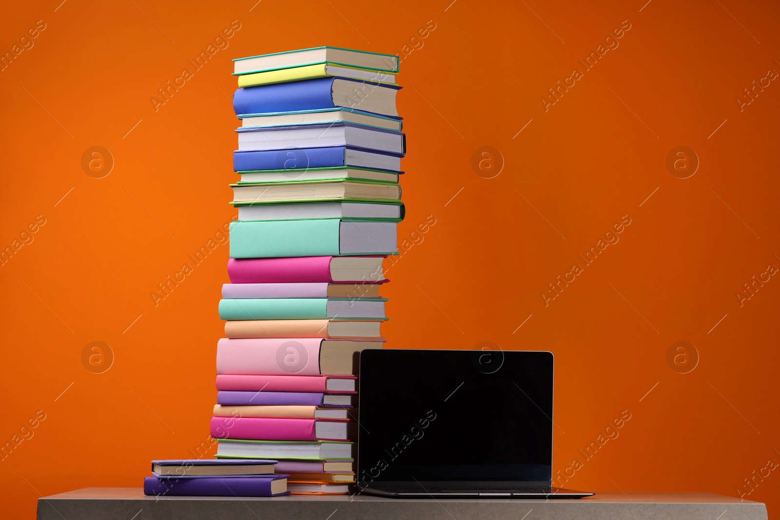 Photo of Stack of colorful books and laptop on wooden table against orange background