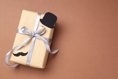 Photo of Happy Father's Day. Gift box with paper hat and moustache on brown background, top view