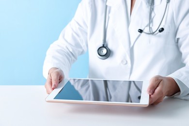 Doctor with tablet at table against light blue background, closeup view