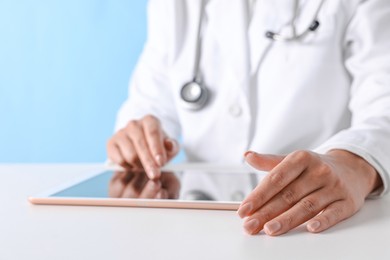 Doctor with tablet at table against light blue background, closeup view