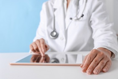 Doctor with tablet at table against light blue background, closeup view