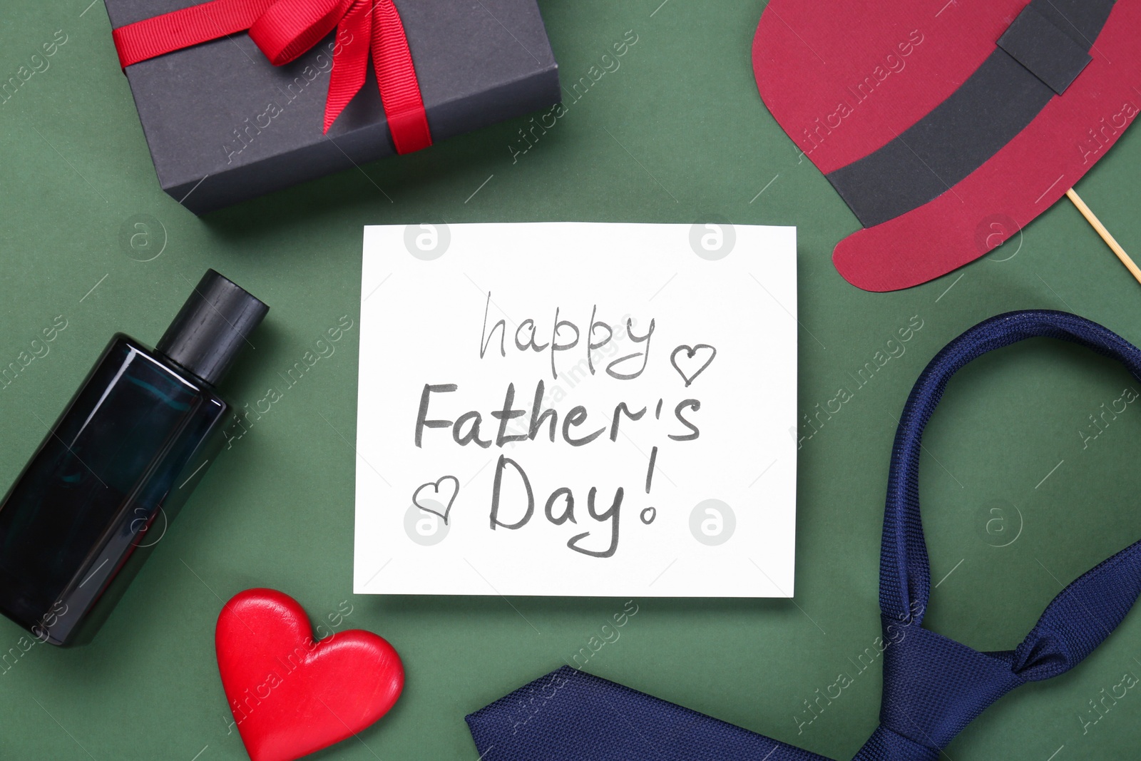 Photo of Card with phrase Happy Father's Day, heart, gifts and paper hat on green background, flat lay