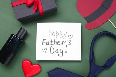 Photo of Card with phrase Happy Father's Day, heart, gifts and paper hat on green background, flat lay