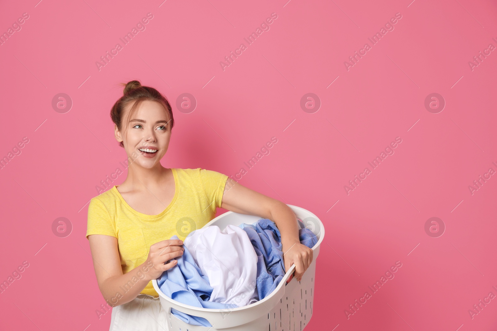 Photo of Happy young housewife with basket full of laundry on pale pink background. Space for text