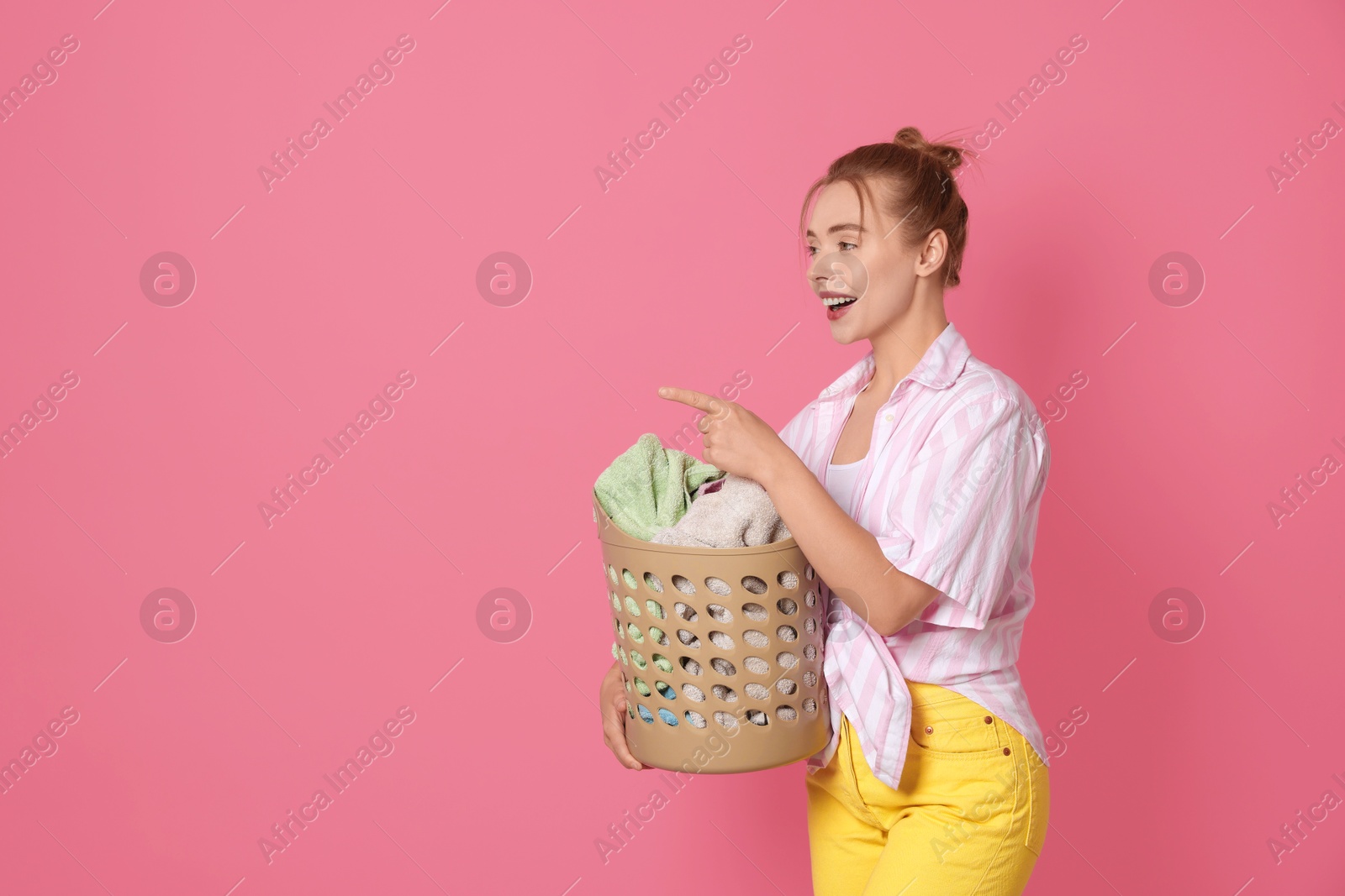 Photo of Happy young housewife with basket full of laundry on pale pink background. Space for text