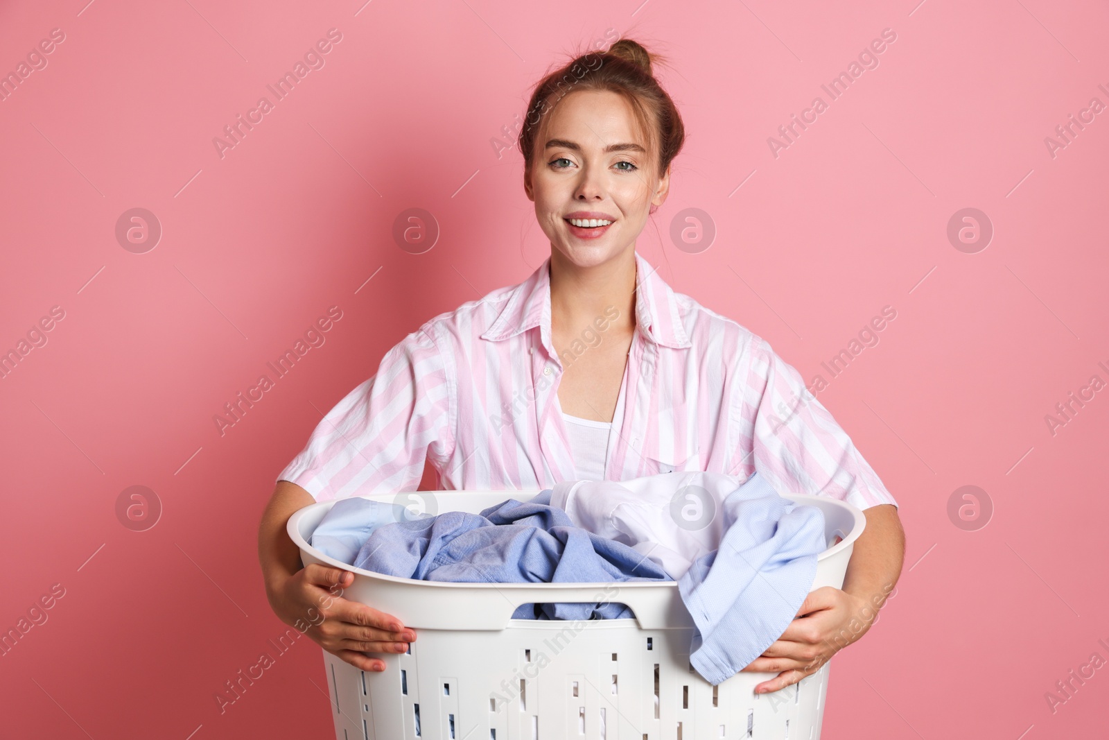 Photo of Happy young housewife with basket full of laundry on pale pink background