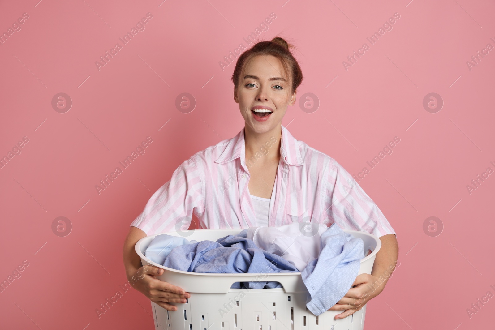 Photo of Happy young housewife with basket full of laundry on pale pink background