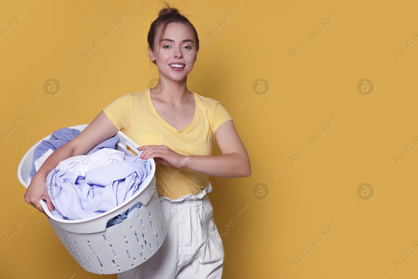 Photo of Happy young housewife with basket full of laundry on yellow background. Space for text