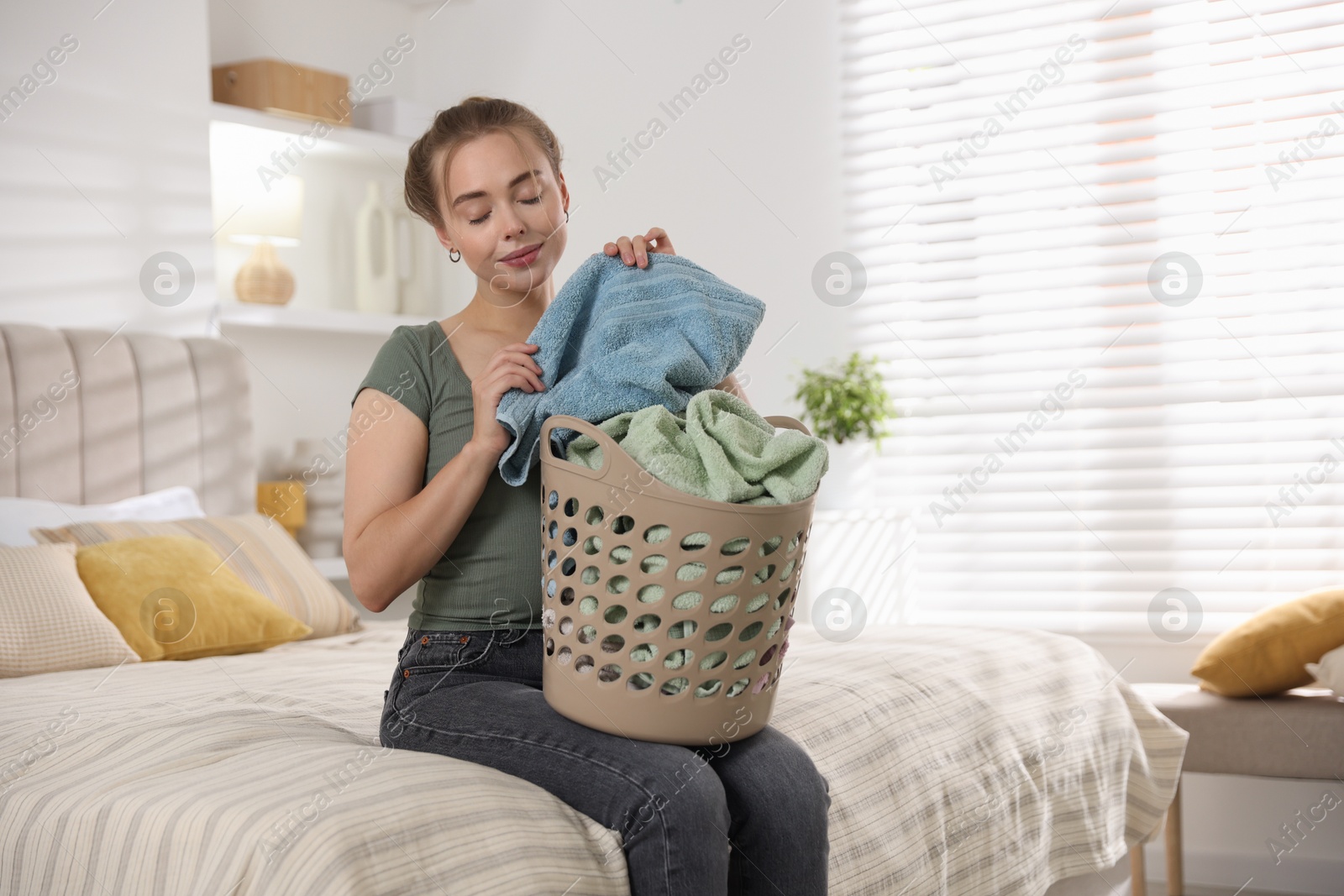 Photo of Happy young housewife with basket full of laundry on bed at home