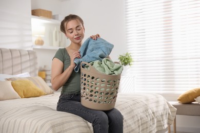 Happy young housewife with basket full of laundry on bed at home