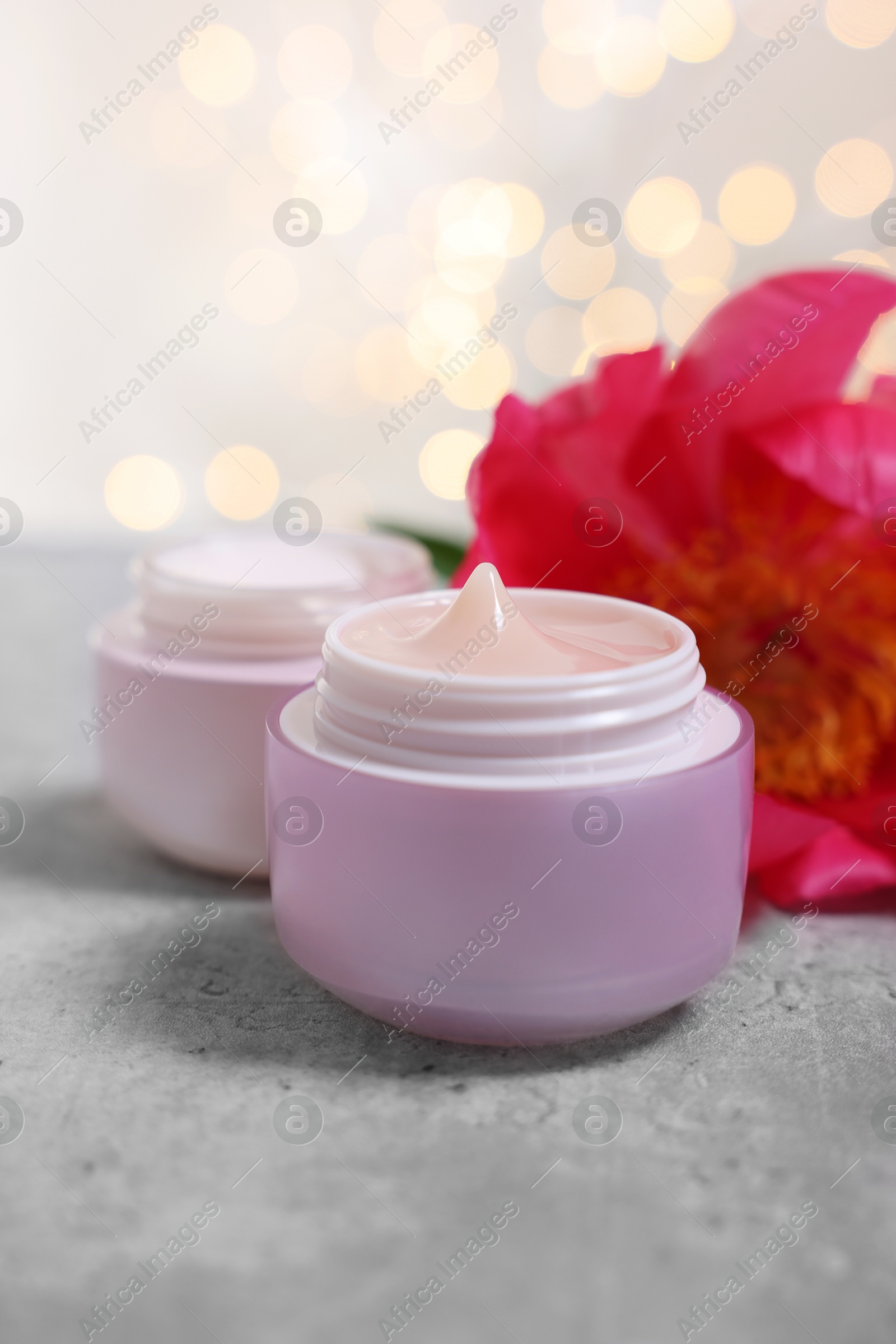 Photo of Jars of creams and peony flower on gray table against light background with blurred lights