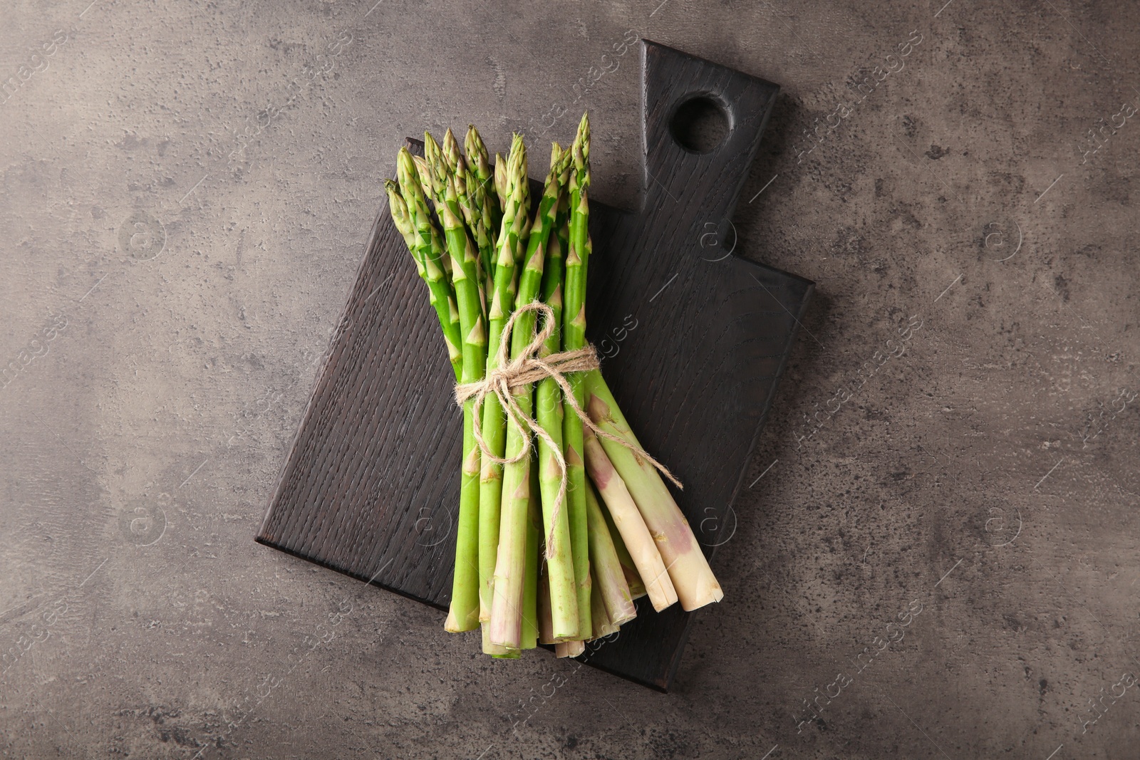 Photo of Board with bunch of fresh green asparagus stems on grey textured table, top view