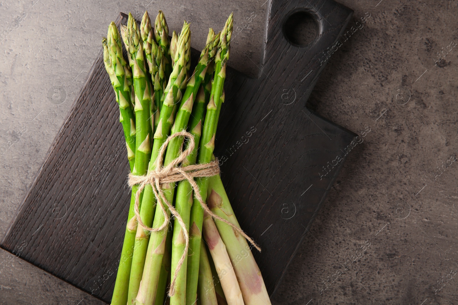 Photo of Board with bunch of fresh green asparagus stems on grey textured table, top view