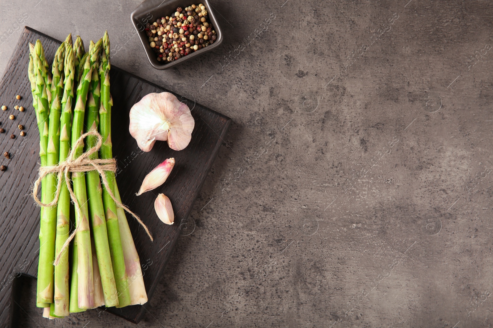 Photo of Board with bunch of fresh green asparagus stems, garlic and peppercorns on grey textured table, flat lay. Space for text