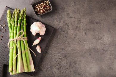 Board with bunch of fresh green asparagus stems, garlic and peppercorns on grey textured table, flat lay. Space for text