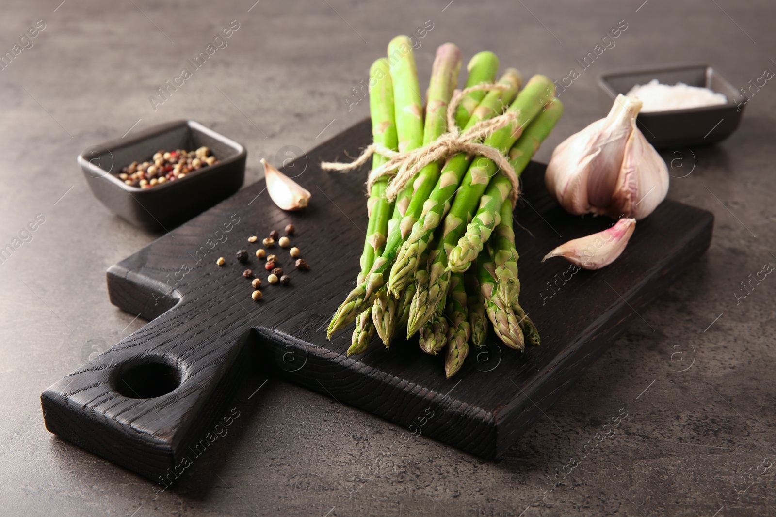 Photo of Board with bunch of fresh green asparagus stems, garlic and peppercorns on grey textured table, closeup