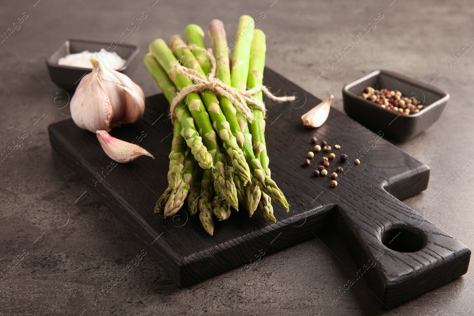 Photo of Board with bunch of fresh green asparagus stems, garlic and peppercorns on grey textured table, closeup