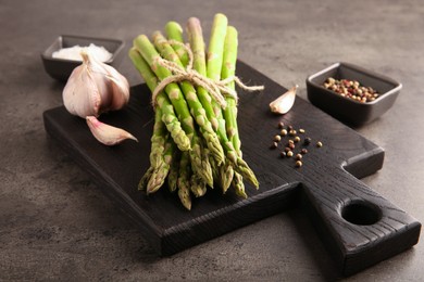 Board with bunch of fresh green asparagus stems, garlic and peppercorns on grey textured table, closeup