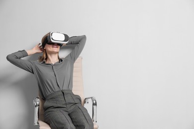 Happy woman with virtual reality headset sitting on chair against gray background, space for text