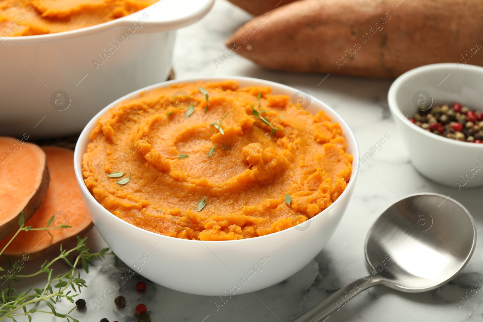 Photo of Delicious mashed sweet potatoes in bowl, spoon, spices and cut vegetable on white marble table, closeup