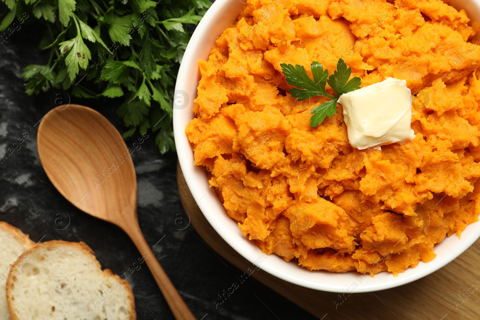 Photo of Delicious mashed sweet potatoes with butter, parsley, bread and spoon on dark table, top view