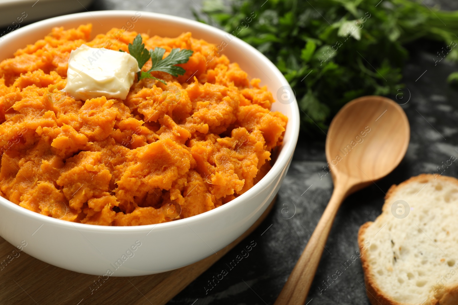 Photo of Delicious mashed sweet potatoes with butter, parsley, bread and spoon on dark table, closeup