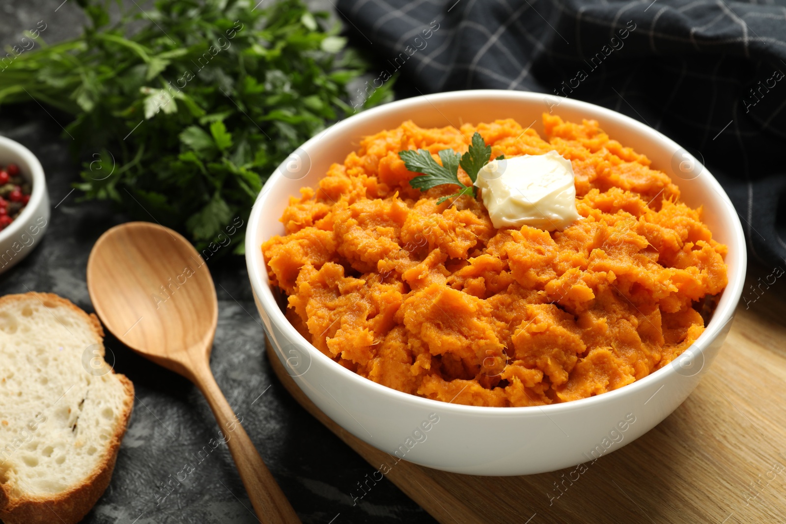 Photo of Delicious mashed sweet potatoes with butter, parsley, bread and spoon on dark table, closeup