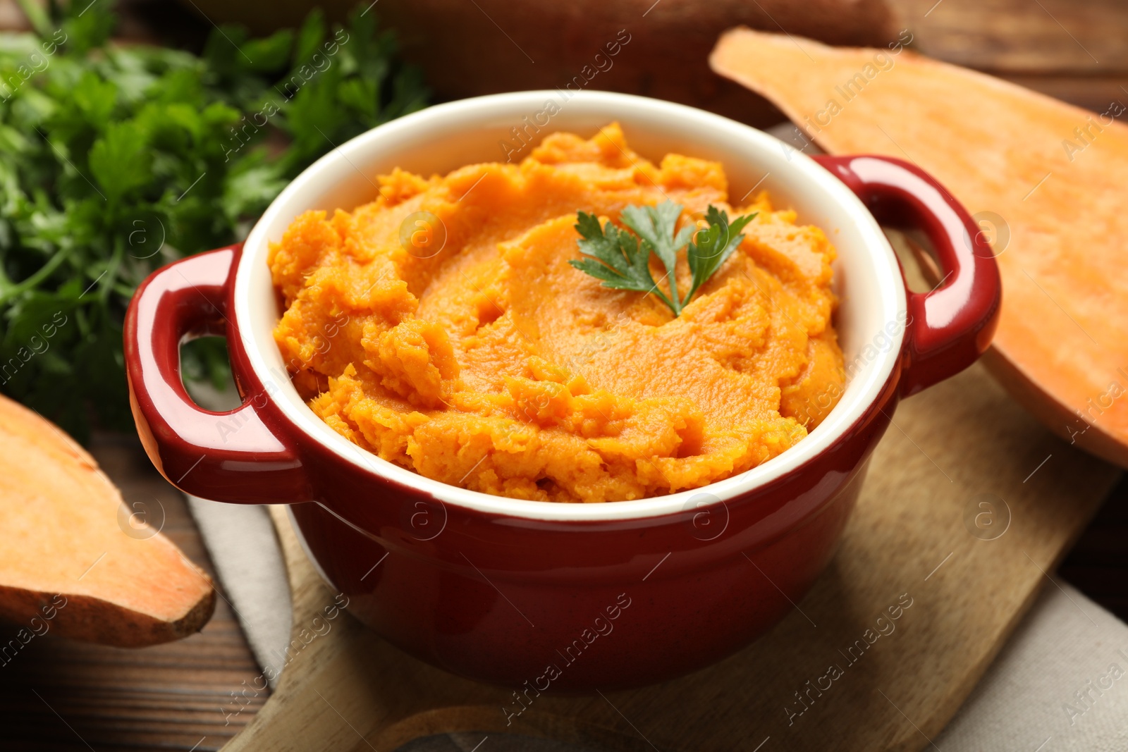 Photo of Delicious mashed sweet potatoes in pot, parsley and cut vegetable on wooden table, closeup