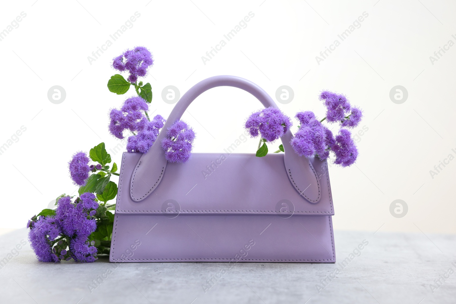 Photo of Stylish violet bag and ageratum flowers on gray table against light background