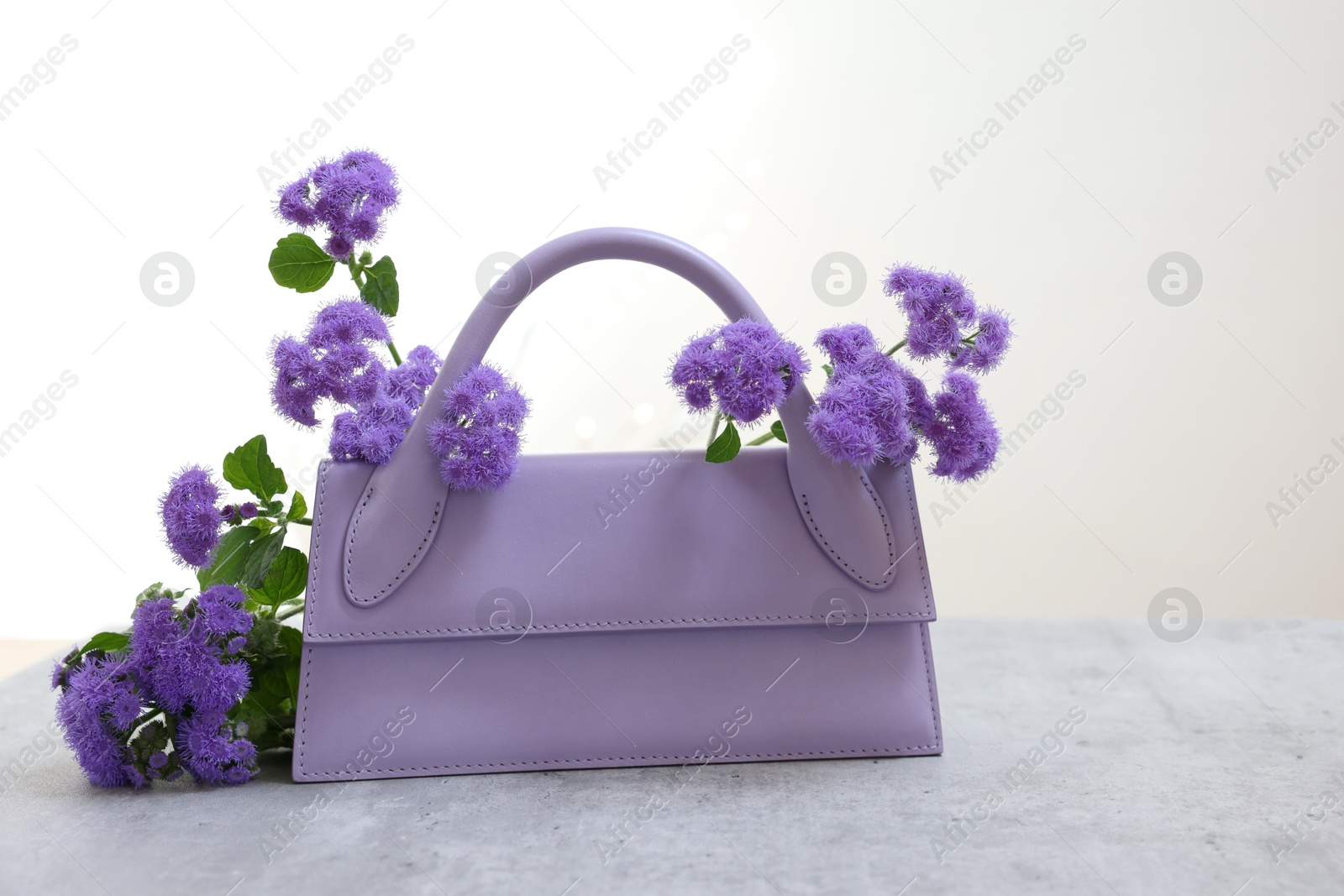 Photo of Stylish violet bag and ageratum flowers on gray table against light background