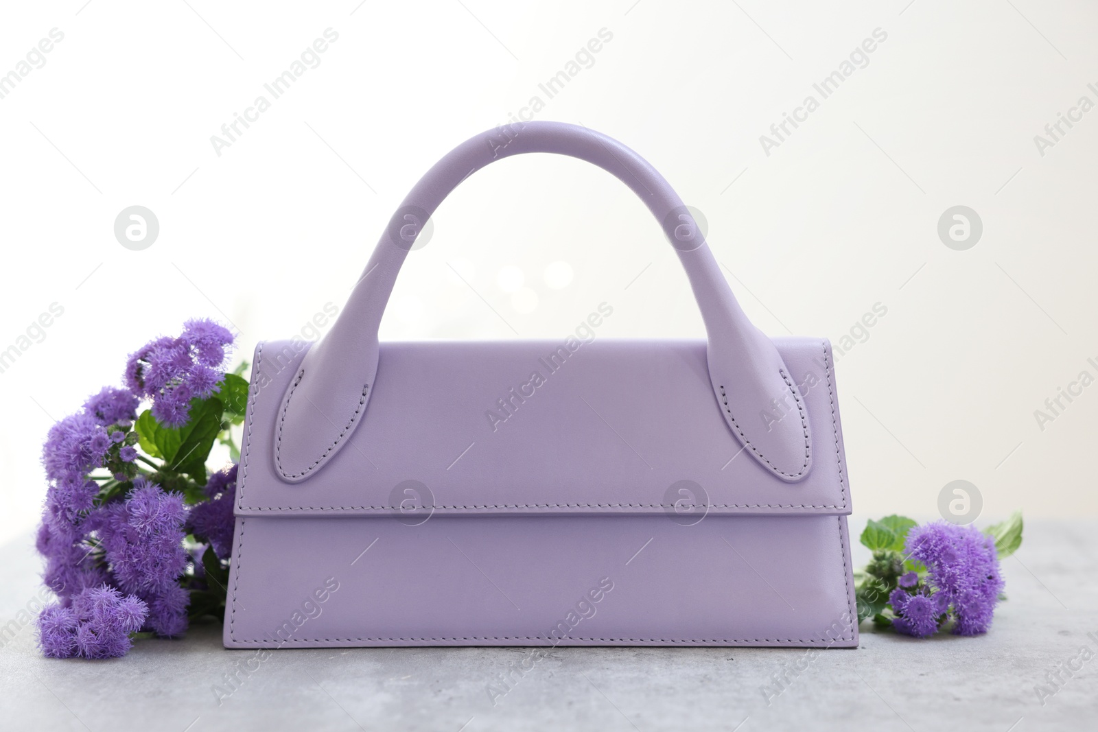 Photo of Stylish violet bag and ageratum flowers on gray table against light background