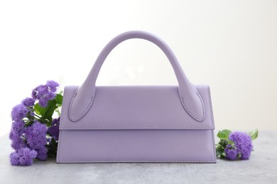 Photo of Stylish violet bag and ageratum flowers on gray table against light background