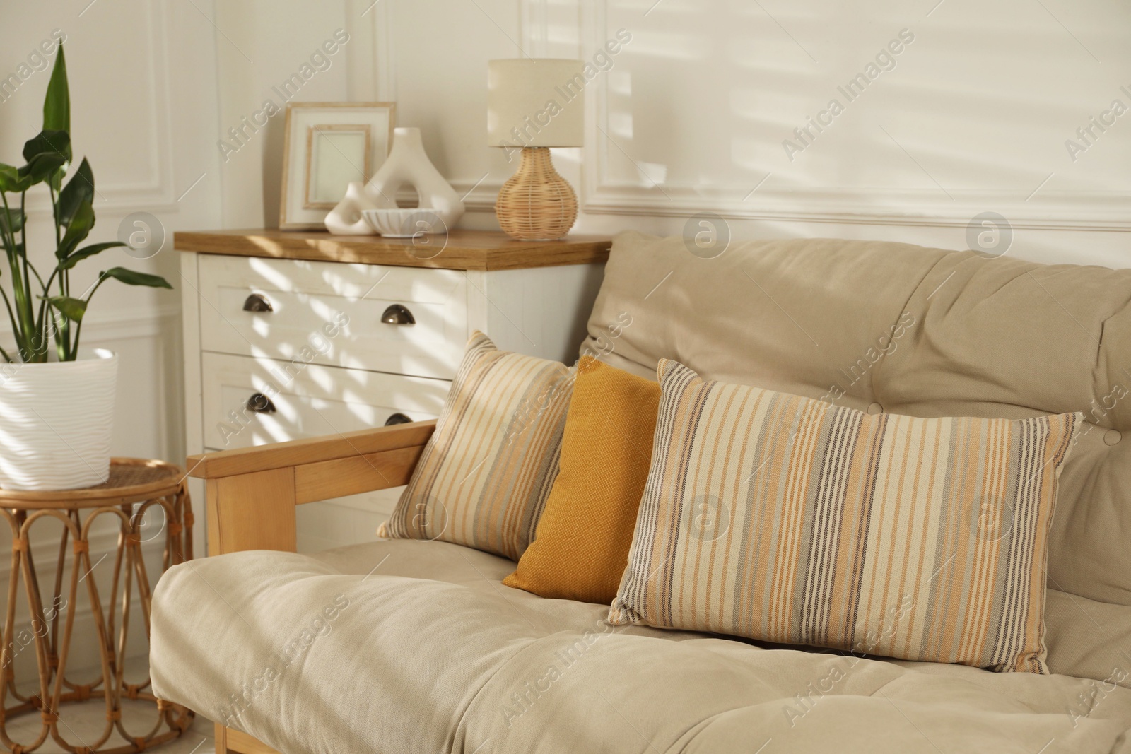 Photo of Sofa with soft pillows, chest of drawers, different decorative elements and houseplant indoors