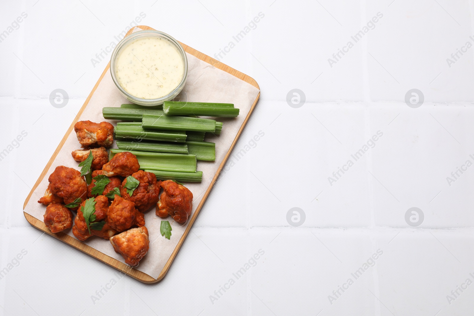 Photo of Baked cauliflower buffalo wings with celery and sauce on white tiled table, top view. Space for text