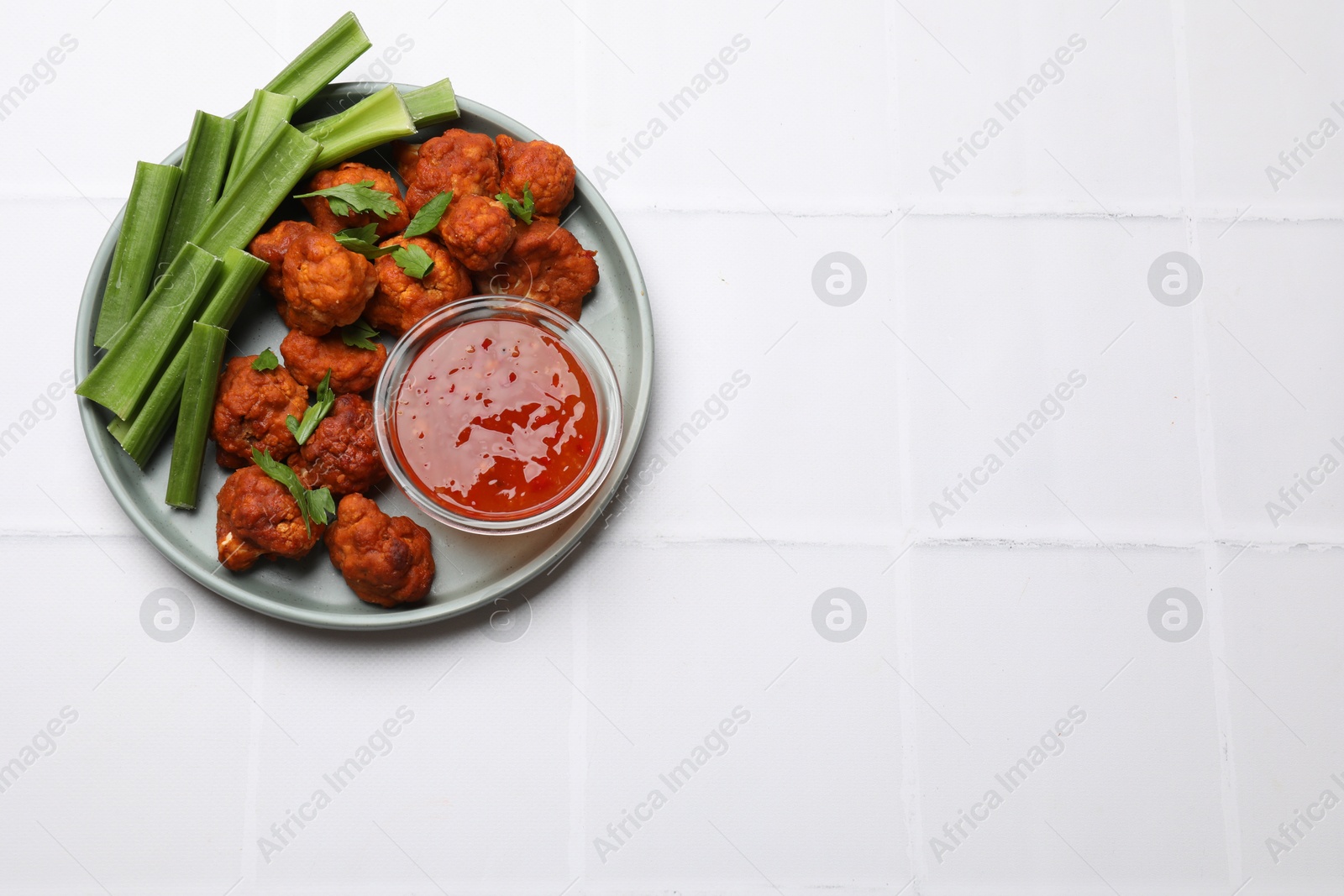 Photo of Baked cauliflower buffalo wings with celery and sauce on white tiled table, top view. Space for text