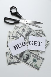 Photo of Cut paper with word Budget, dollar banknotes and scissors on white background, top view