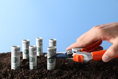 Photo of Budgeting. Man cutting dollar banknotes with scissors on soil against light blue background, closeup