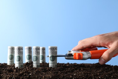 Photo of Budgeting. Man cutting dollar banknotes with scissors on soil against light blue background, closeup