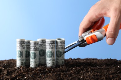 Photo of Budgeting. Man cutting dollar banknotes with scissors on soil against light blue background, closeup