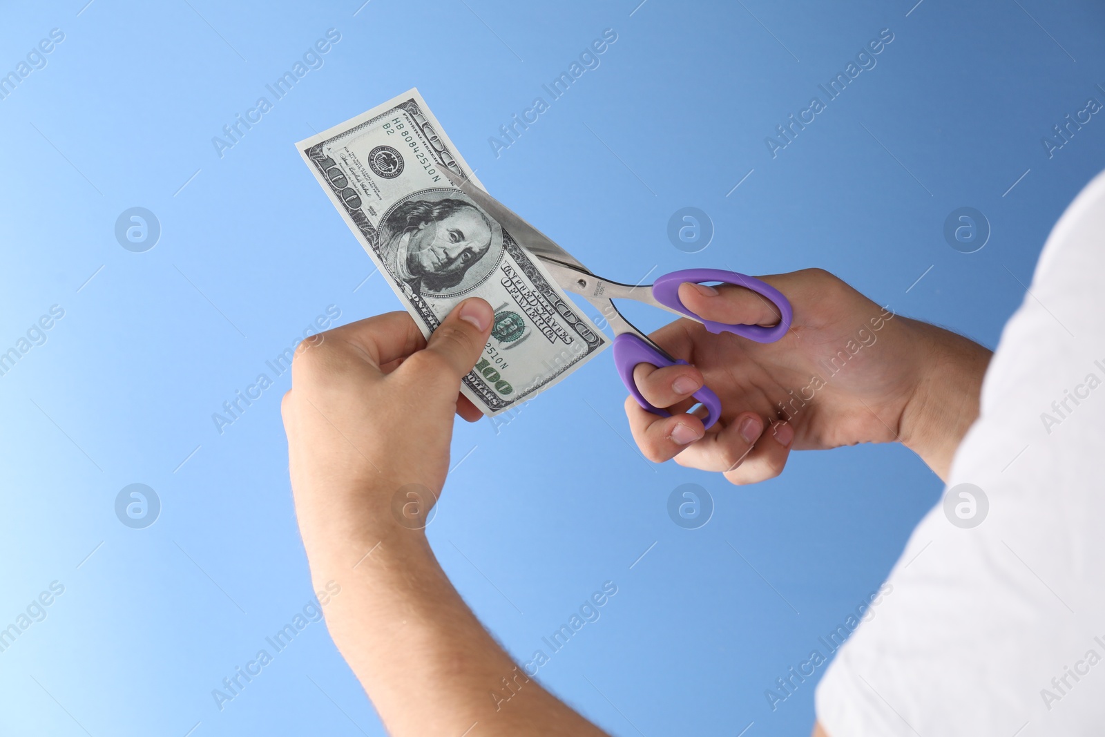 Photo of Budgeting. Man cutting dollar banknote with scissors on light blue background, closeup