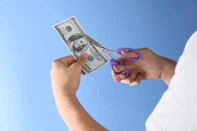 Photo of Budgeting. Man cutting dollar banknote with scissors on light blue background, closeup