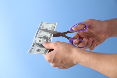 Photo of Budgeting. Man cutting dollar banknote with scissors on light blue background, closeup