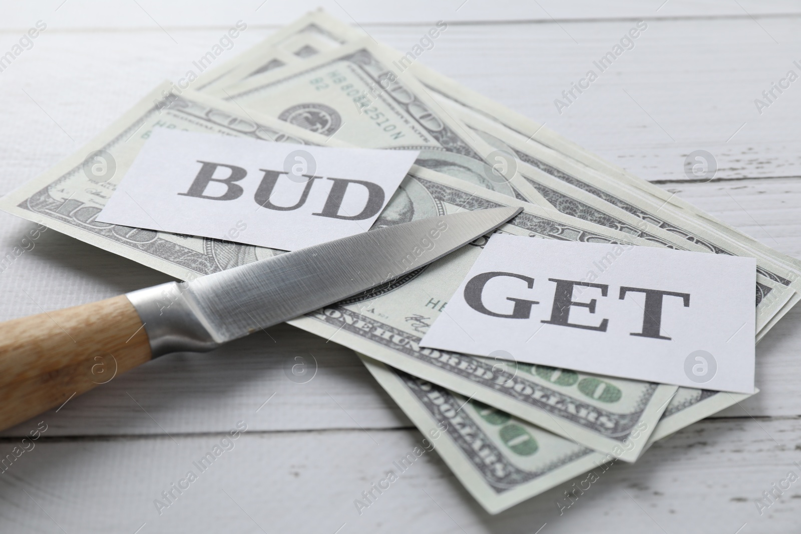 Photo of Cut paper with word Budget, dollar banknotes and knife on white wooden table, closeup