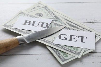 Cut paper with word Budget, dollar banknotes and knife on white wooden table, closeup