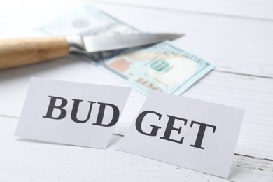 Photo of Cut paper with word Budget, dollar banknote and knife on white wooden table, closeup